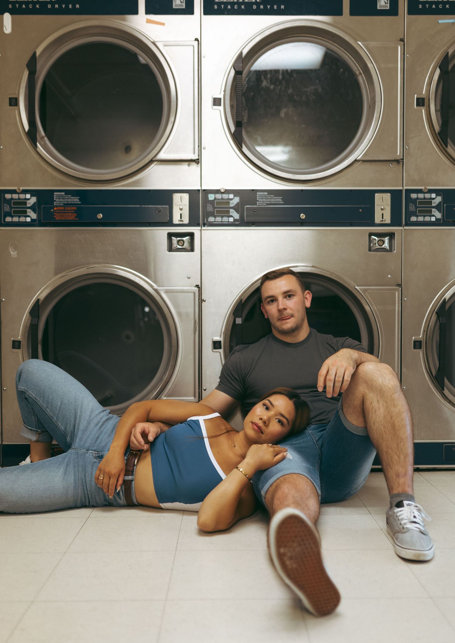 Relaxed couple posing in front of stacked industrial dryers on the floor of a laundromat.