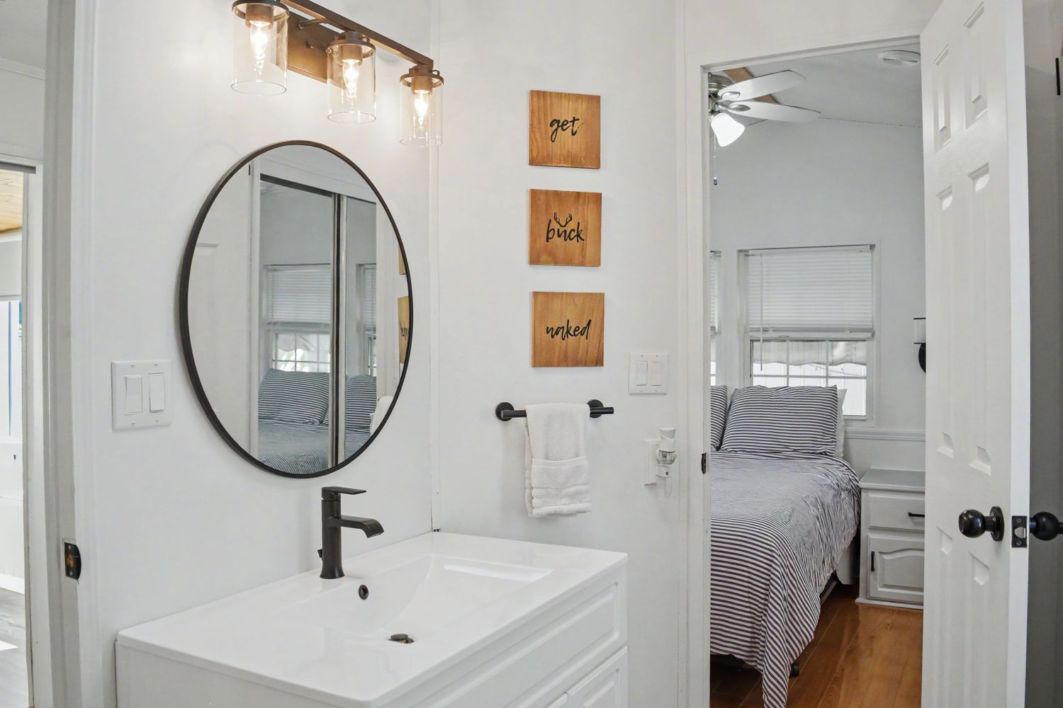 Modern bathroom with round mirror, white vanity, black fixtures, and view into bedroom with natural light.