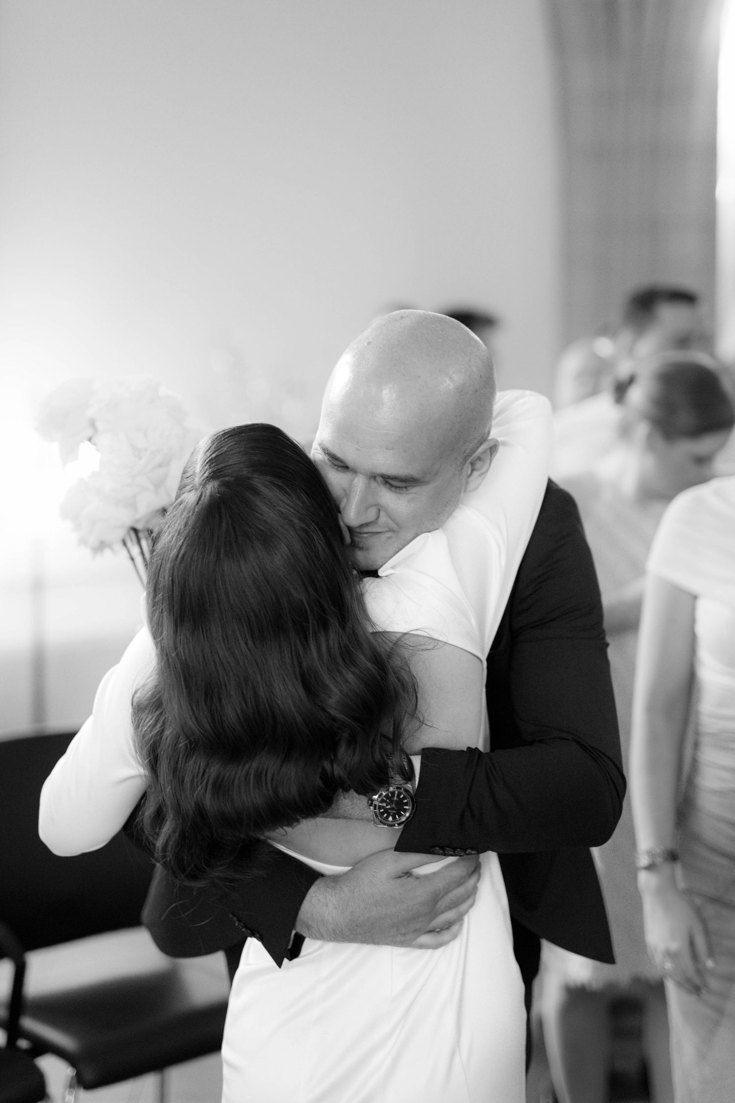 Black and white photo of a bald groom embracing bride holding white flowers at a wedding ceremony.