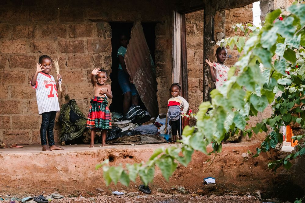 Four young children standing outside a rustic brick building in a rural African village setting.