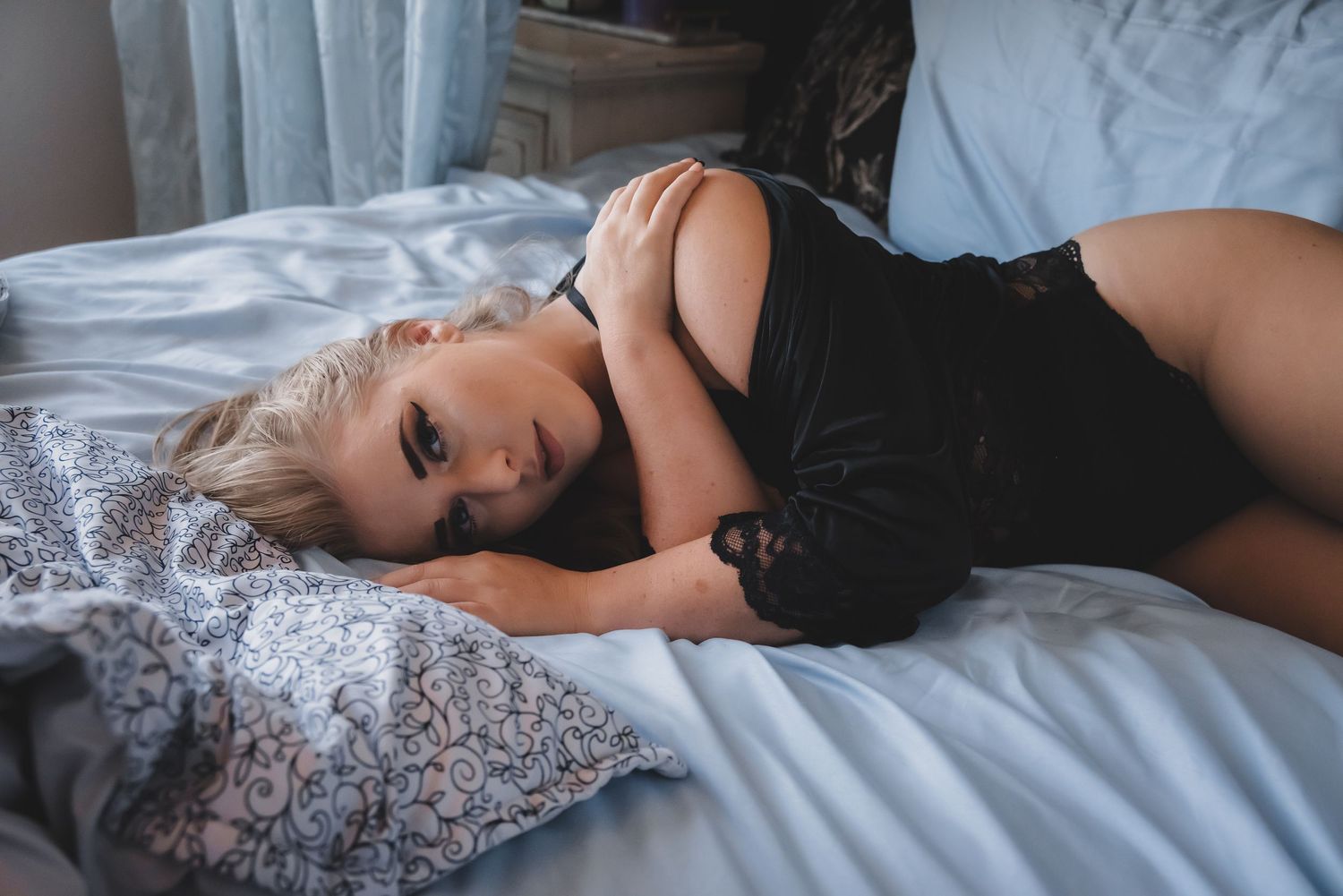 Blonde woman in black lingerie poses on bed with blue sheets.