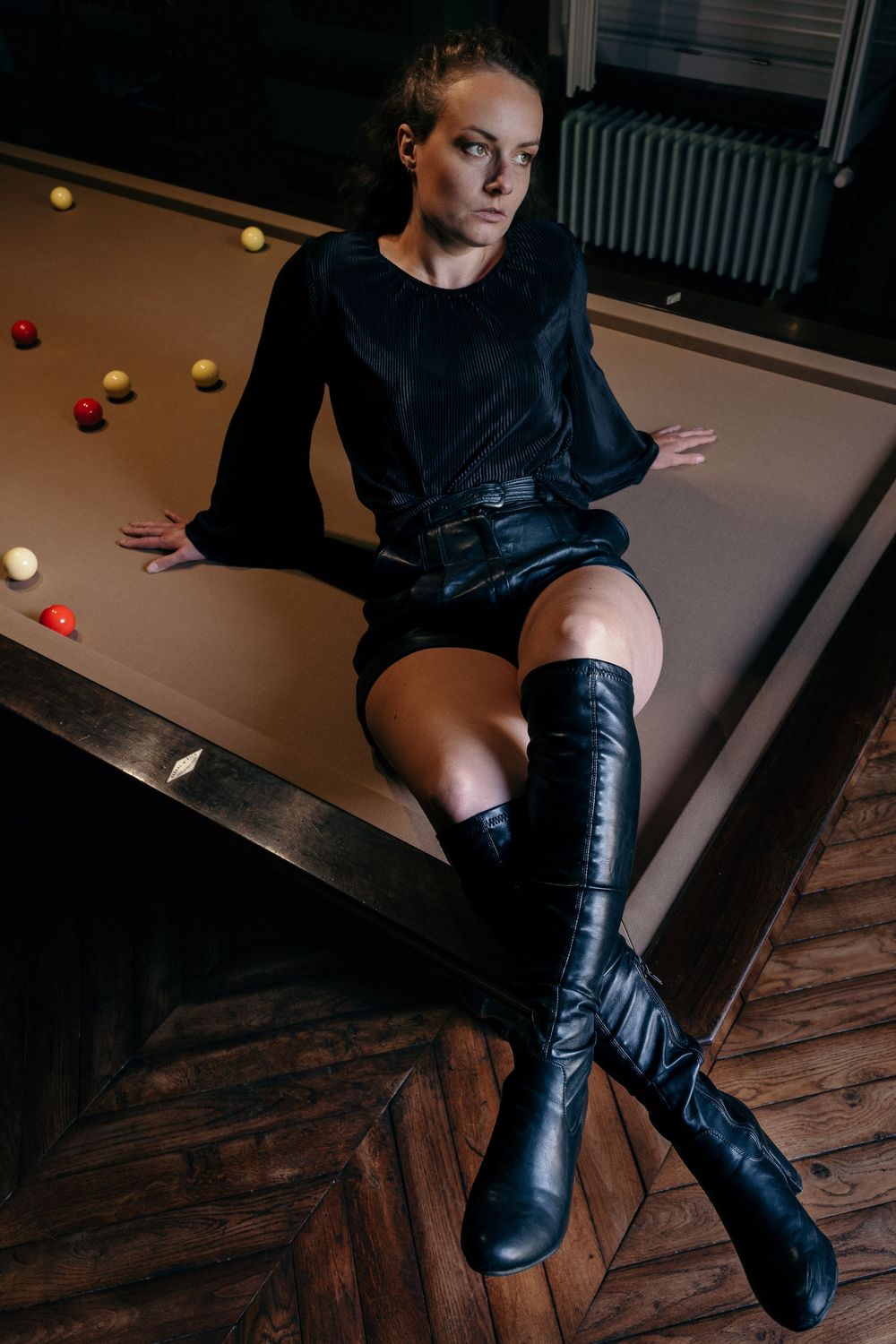 Portrait d’une femme en tenue noire assise sur un billard dans une ambiance intérieure cinématographique.