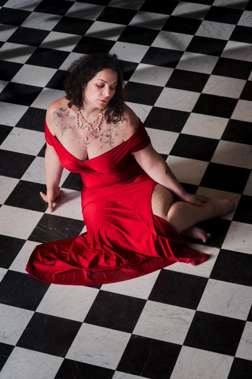 Portrait en plongée d’une femme en robe rouge assise sur un sol en damier noir et blanc.