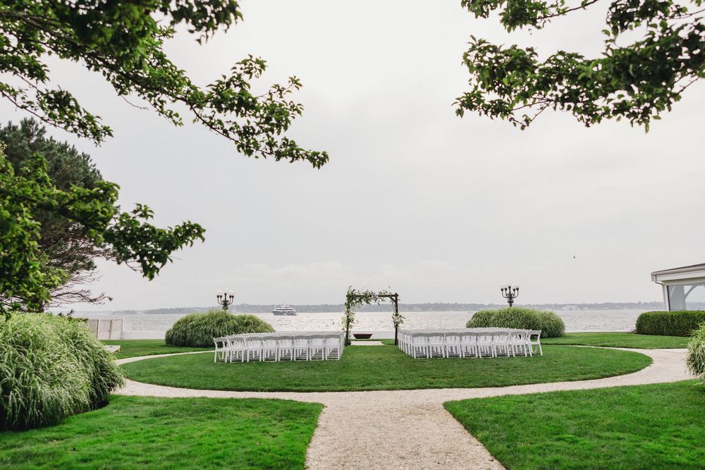 Elegant outdoor wedding ceremony setup on lush green lawn overlooking serene waterfront with white chairs and floral arch.