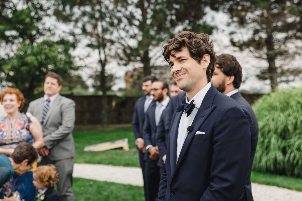 Smiling groom in navy suit and bow tie reacts emotionally at outdoor wedding ceremony with guests in background.
