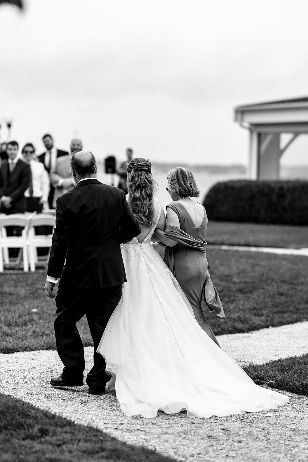 Black and white photo of a bride being walked down the aisle by her parents at an outdoor wedding ceremony.