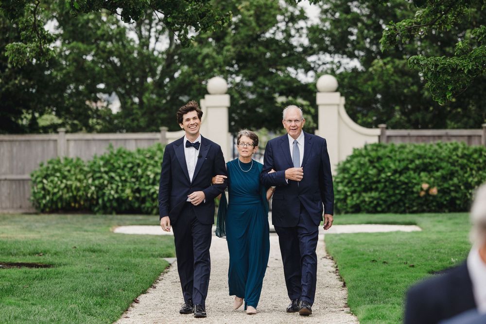 Groom walks with parents down garden path at outdoor wedding ceremony in formal attire.
