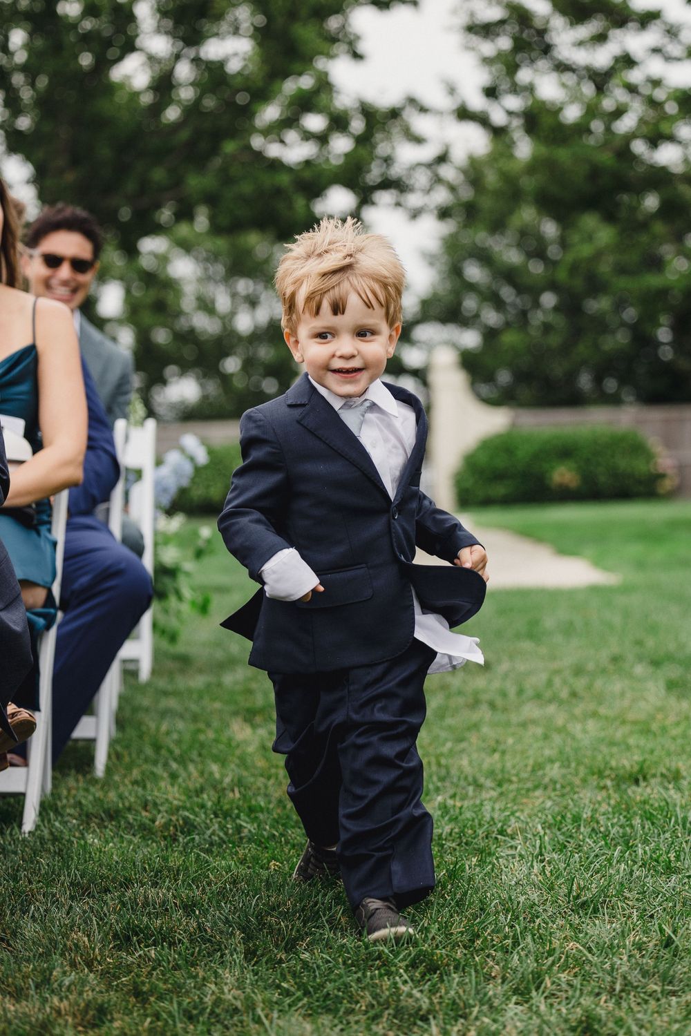 Adorable ring bearer in navy suit walks down outdoor wedding aisle on green grass, smiling joyfully.
