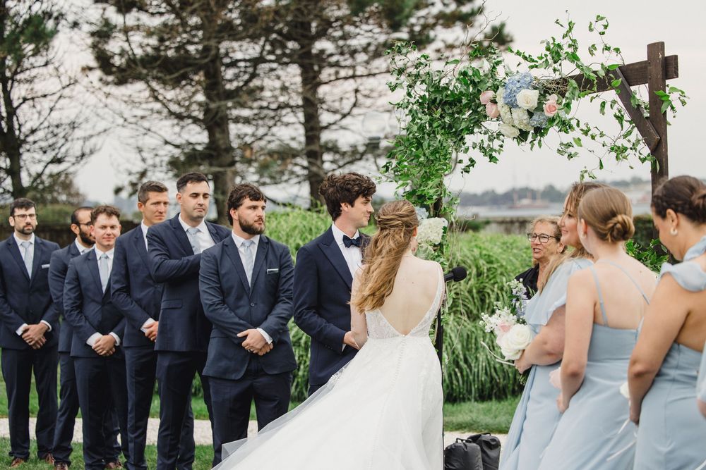 Outdoor wedding ceremony with groom in navy suit, groomsmen, and bridesmaids in blue dresses beside a floral wooden arch.