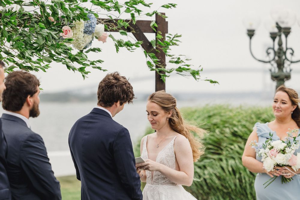 Outdoor wedding ceremony by waterfront with bride and groom exchanging vows under floral arch, bridesmaid holding bouquet.