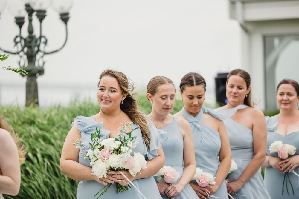 Bridesmaids in light blue dresses holding white and pink bouquets at an outdoor wedding ceremony.