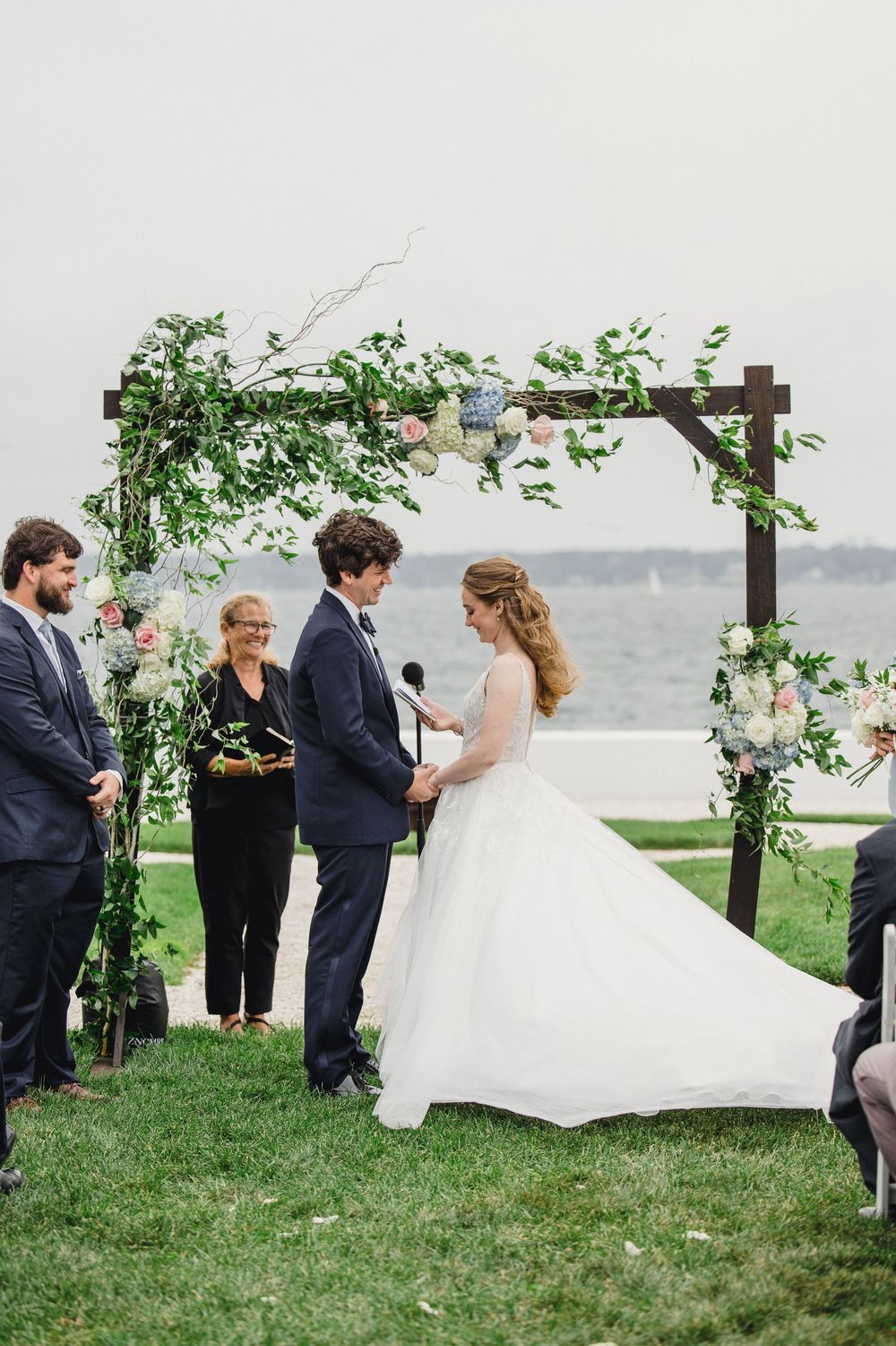 Bride and groom exchange vows under a floral wooden arch by the waterfront during an outdoor wedding ceremony.