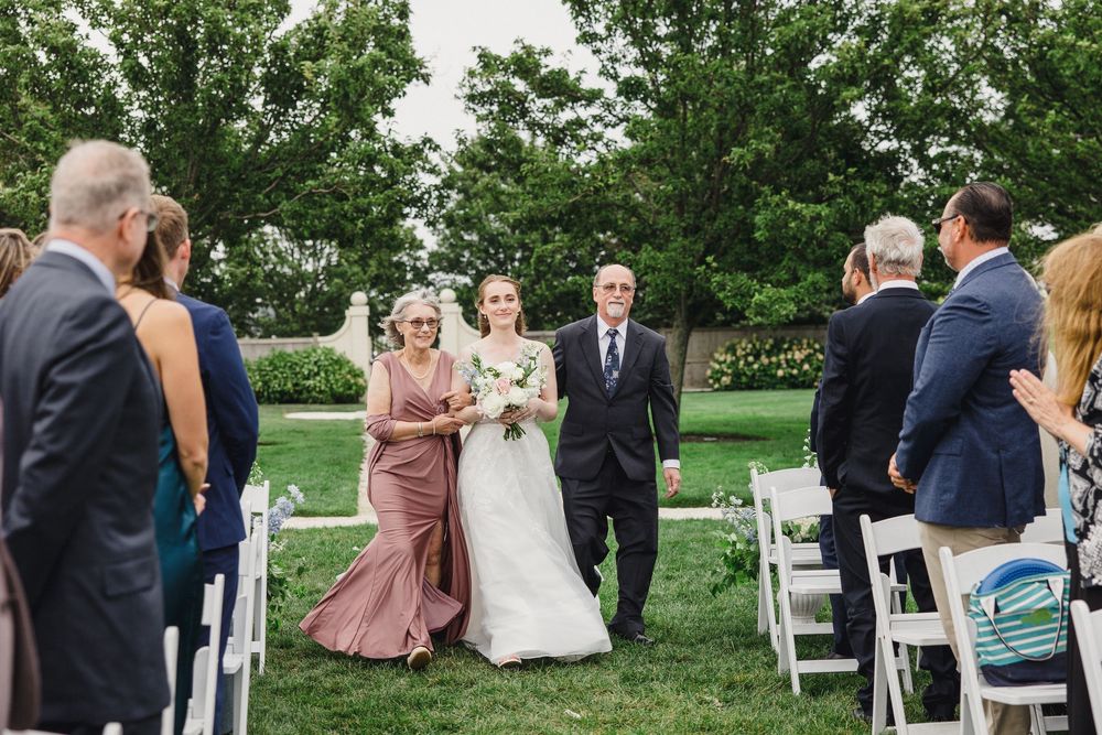 Bride walks down the aisle with parents at an elegant outdoor wedding ceremony with white chairs and lush green trees.