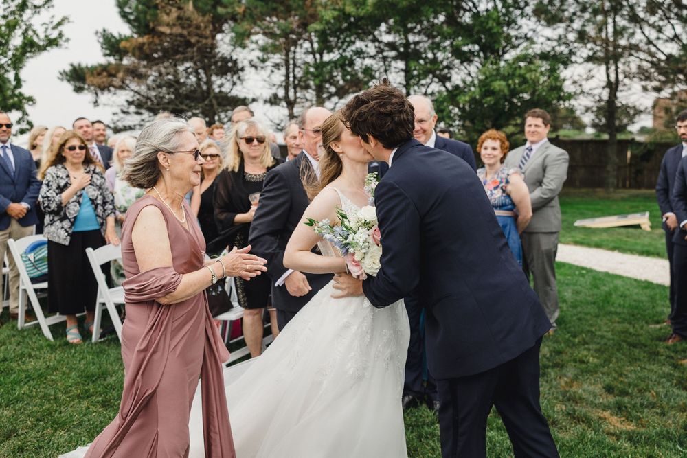 Groom kisses bride on cheek during outdoor wedding ceremony while guests watch and mother of bride applauds.
