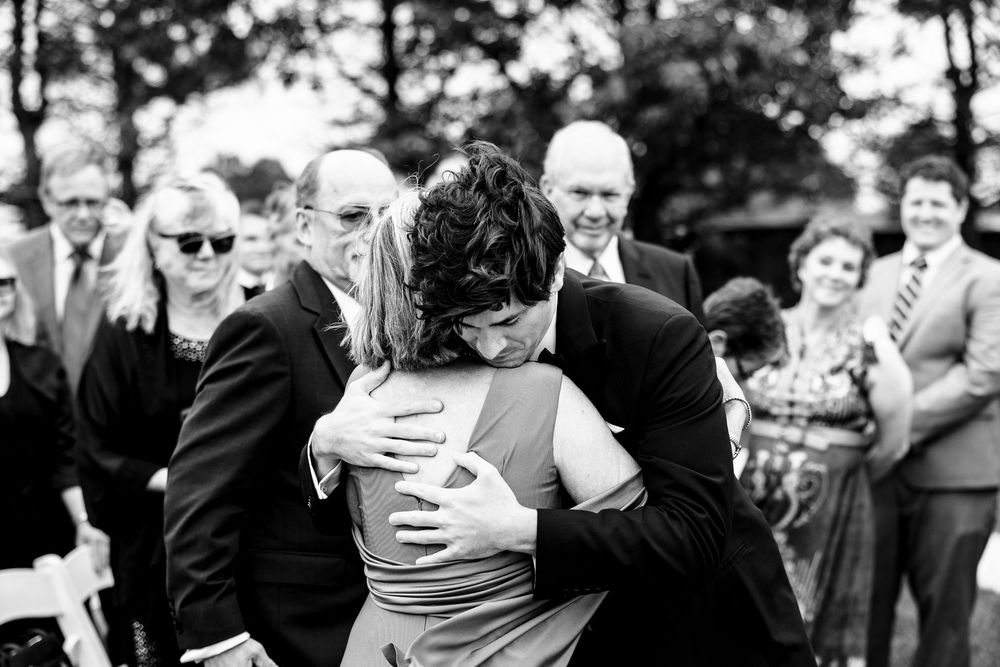 Black and white photo of groom embracing family member at outdoor wedding ceremony with guests watching.
