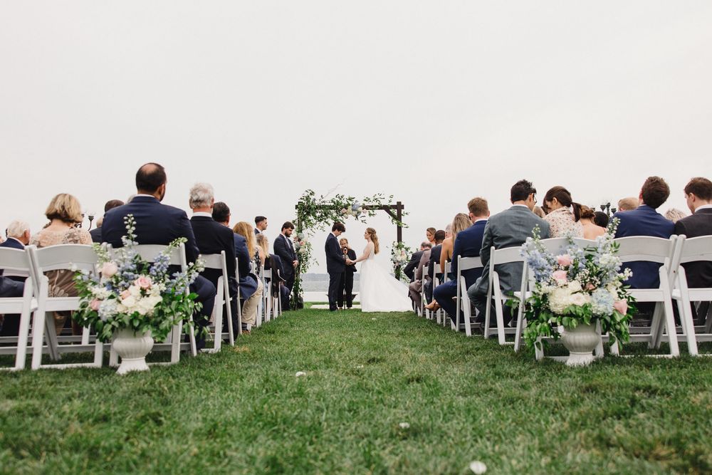 Outdoor wedding ceremony with guests seated in white chairs on green lawn, couple at floral arch altar.