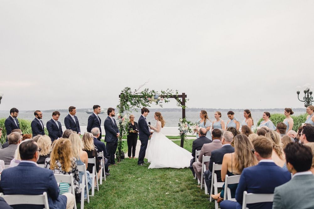 Outdoor wedding ceremony on a waterfront lawn with guests seated, bridal party standing at a floral arch altar.