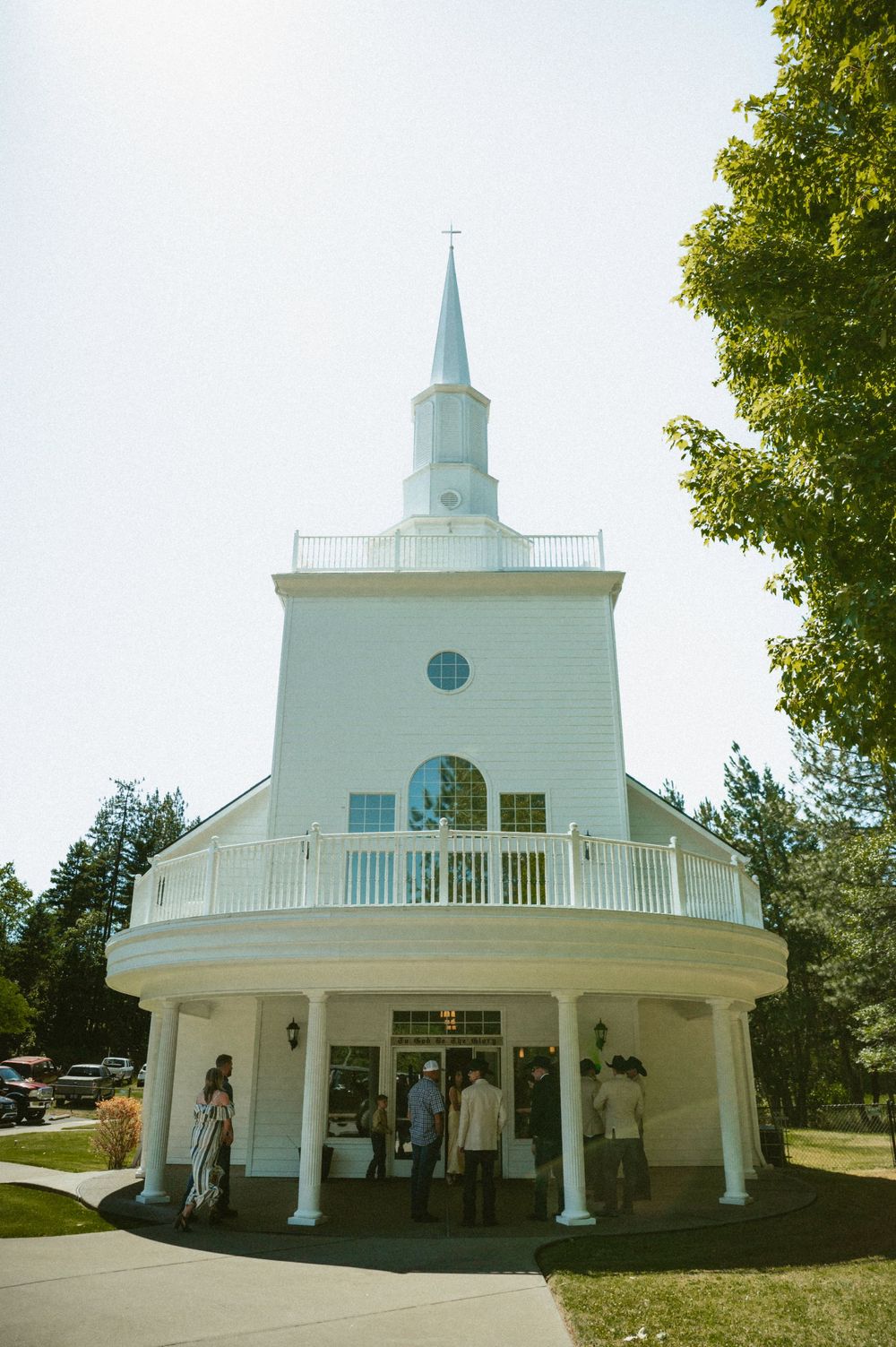 White chapel with steeple and wraparound porch with guests gathering outside on a sunny day.