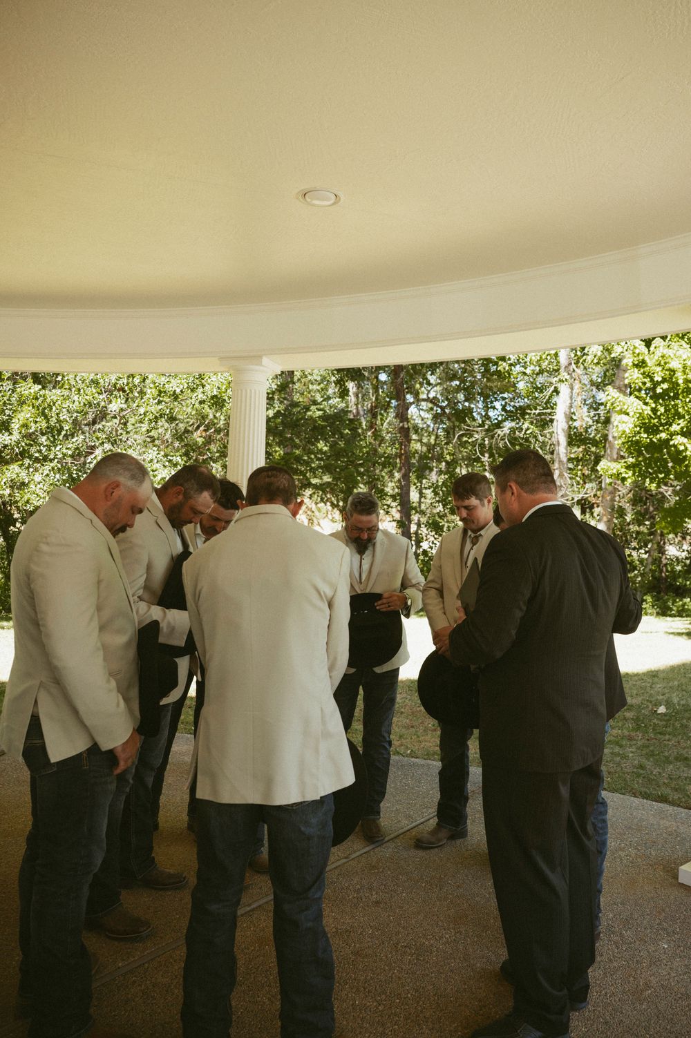 Groomsmen in suits and jeans gather under an outdoor pavilion for a prayer circle before a wedding ceremony.