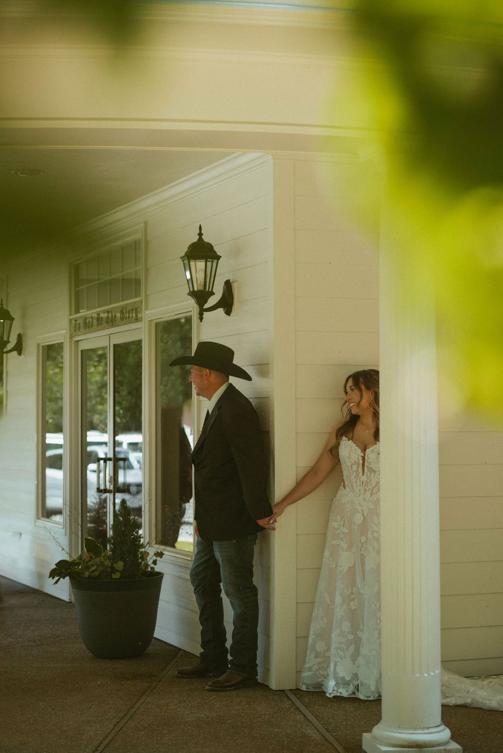 A groom in a black cowboy hat holds hands with a bride in a lace wedding dress on a white porch.