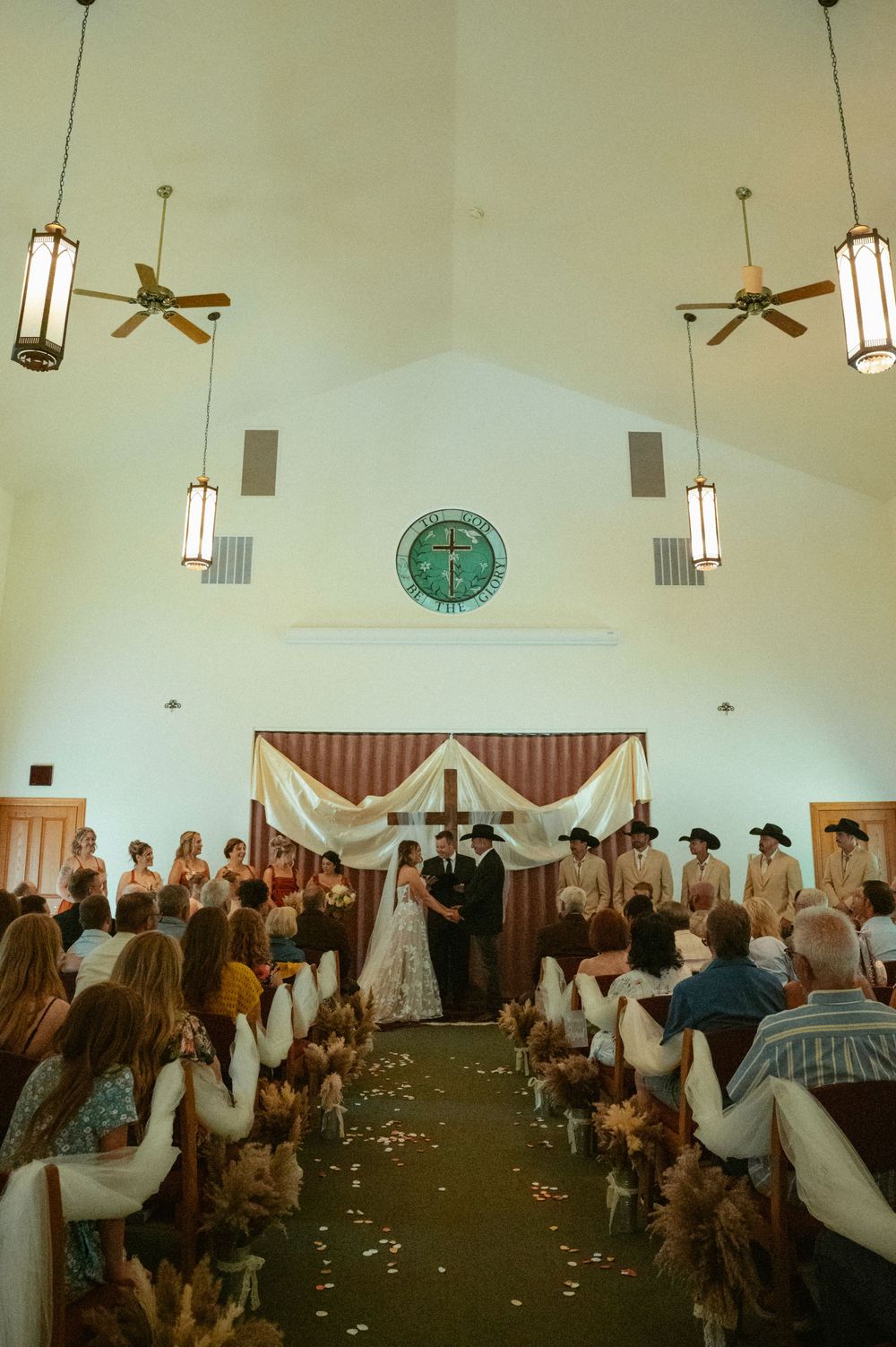 Wedding ceremony in a church with cross, floral aisle, bride and groom at altar, guests seated in pews.