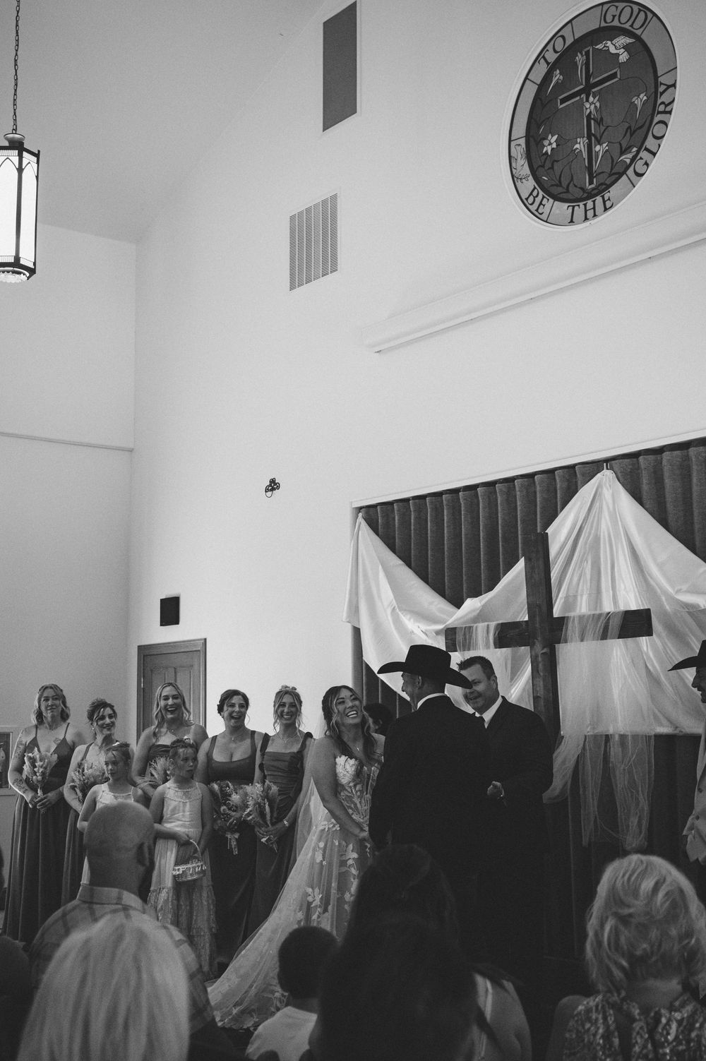 Black and white photo of a wedding ceremony in a church with cross backdrop, bridal party, and guests gathered.