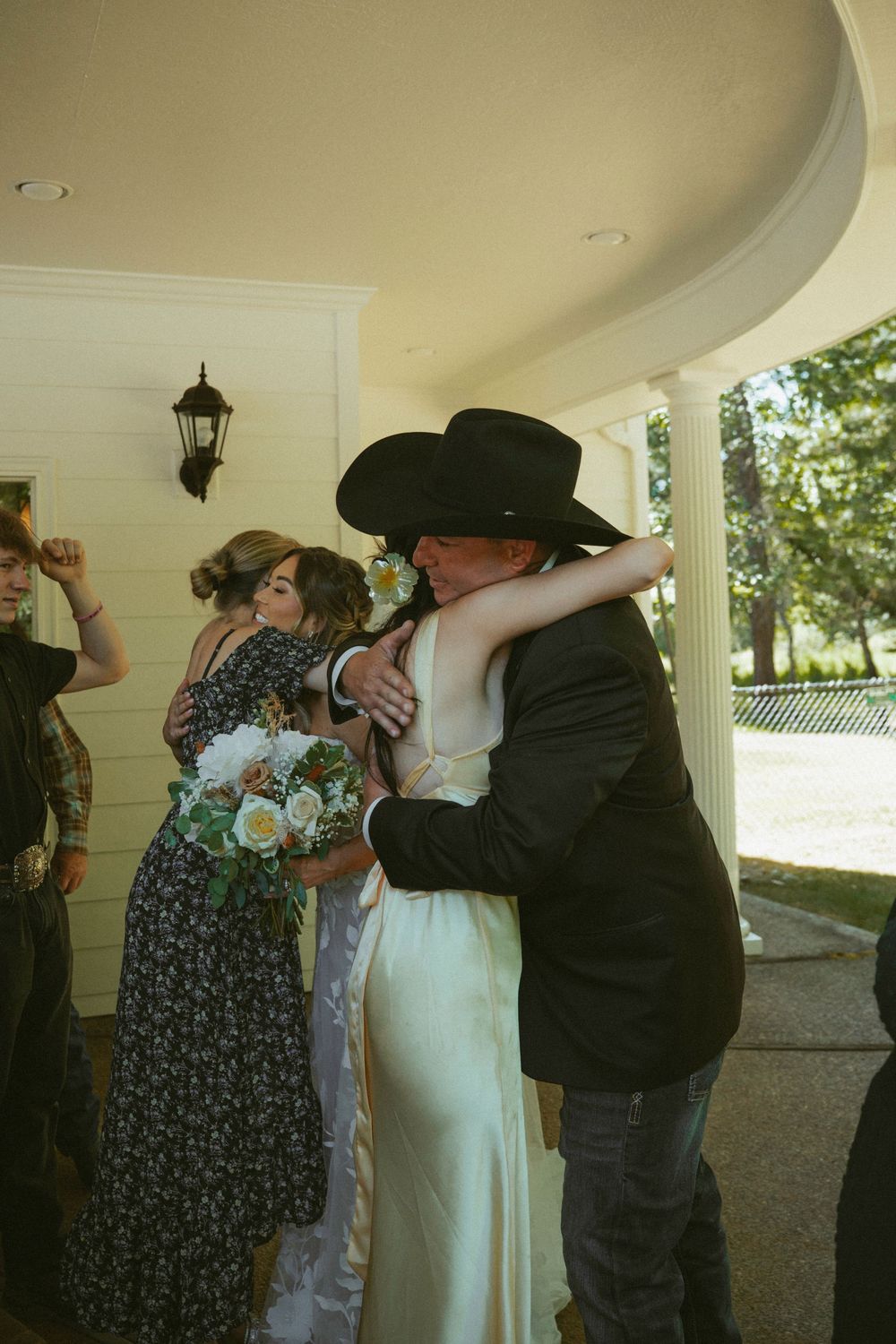 Bride in white gown hugging man in black cowboy hat outside wedding venue.