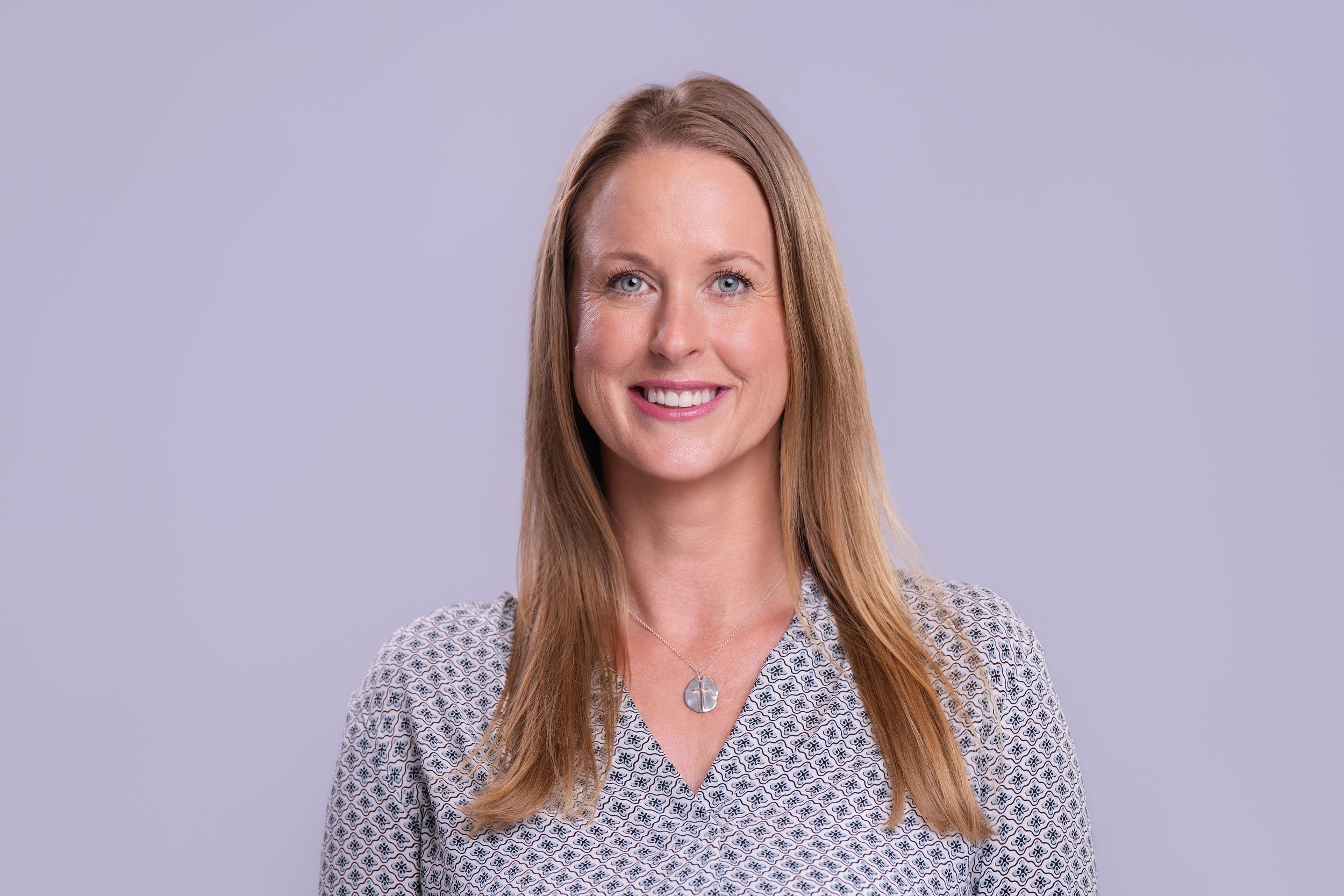 Professional headshot of a smiling blonde woman in a patterned blouse against a gray background.