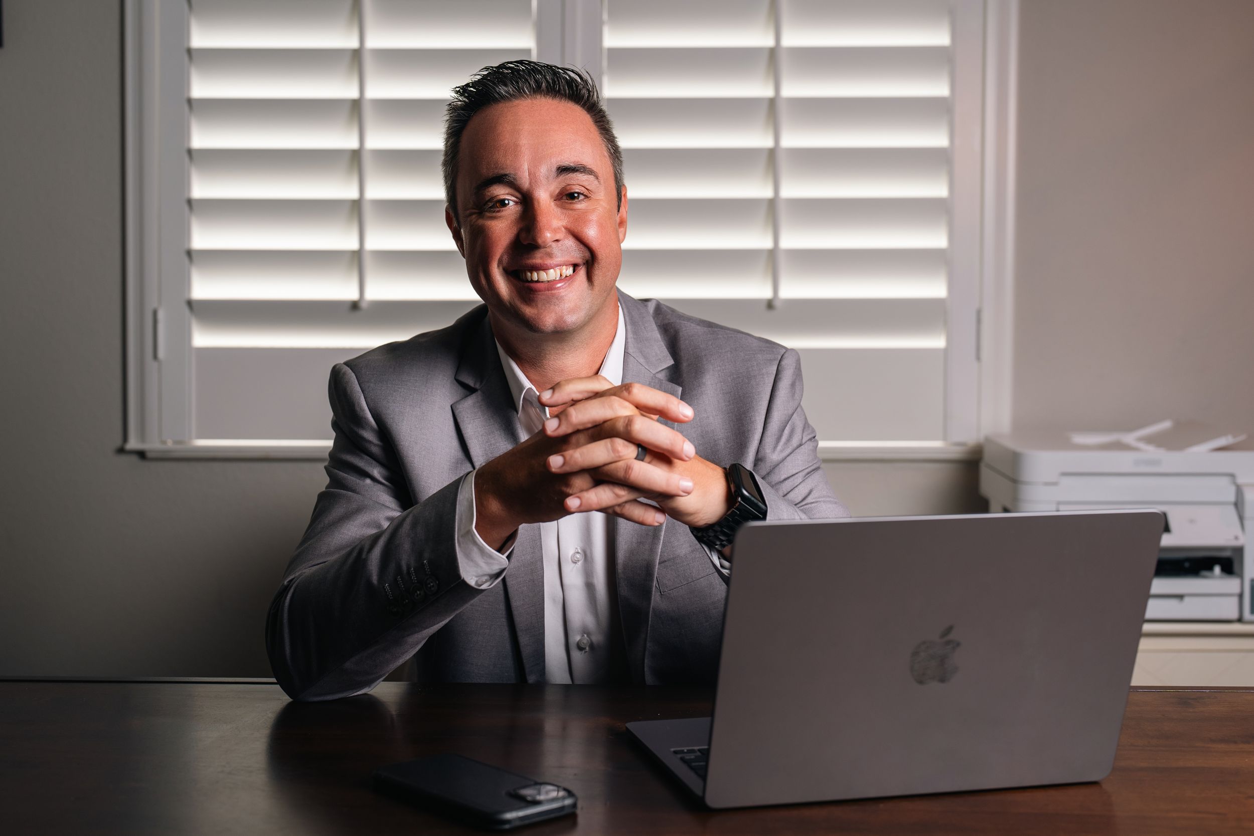 Smiling businessman in gray blazer sitting at desk with MacBook laptop, clasping hands in bright modern home office.
