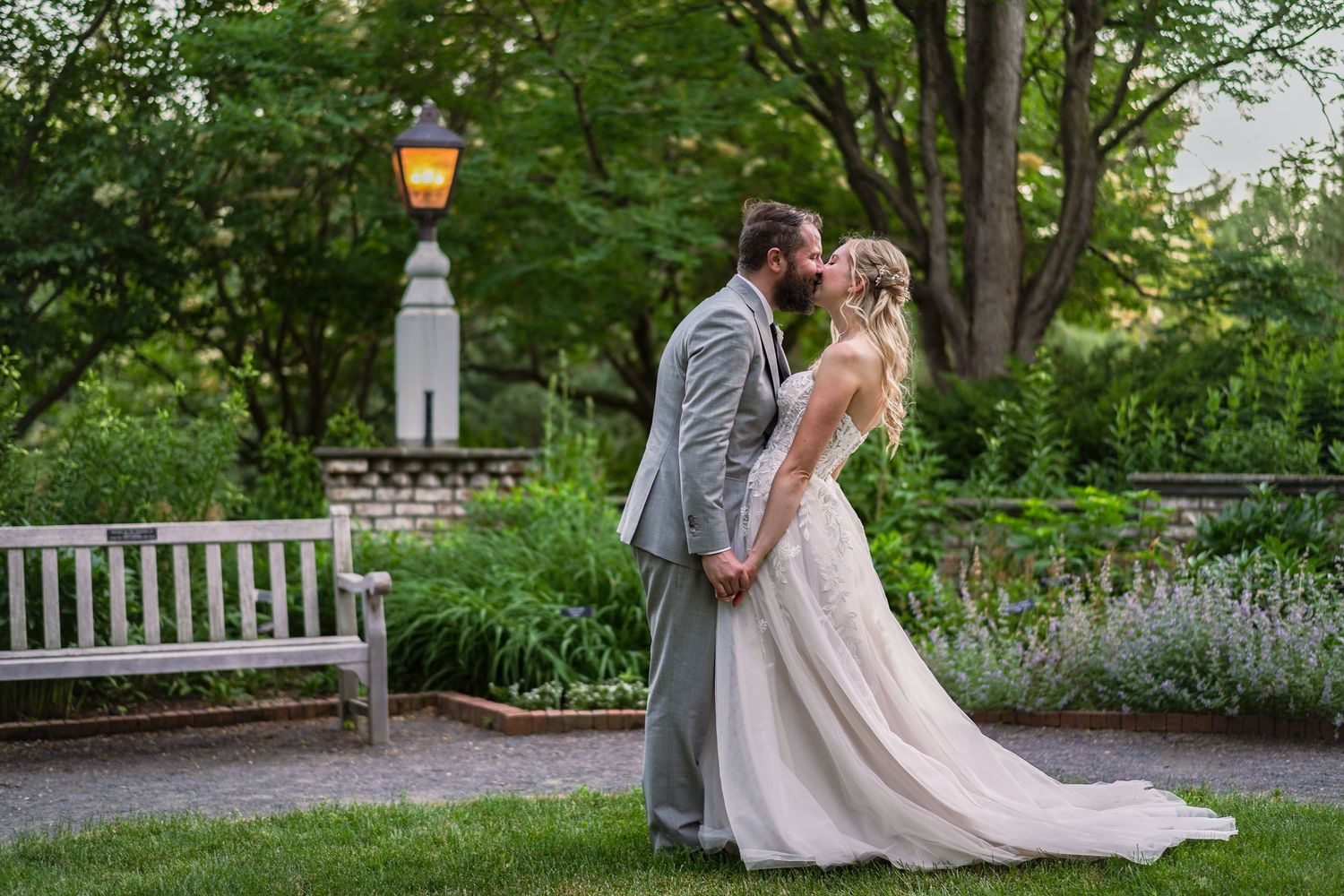 Bride and groom sharing a romantic kiss near a garden bench surrounded by greenery.