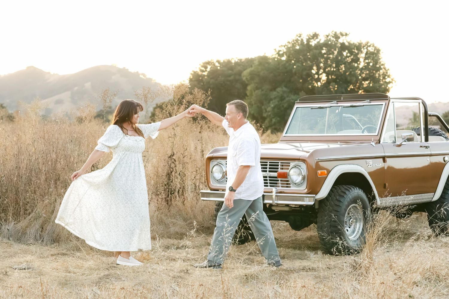 Couple dancing in golden field near vintage Ford Bronco with mountains at sunset.