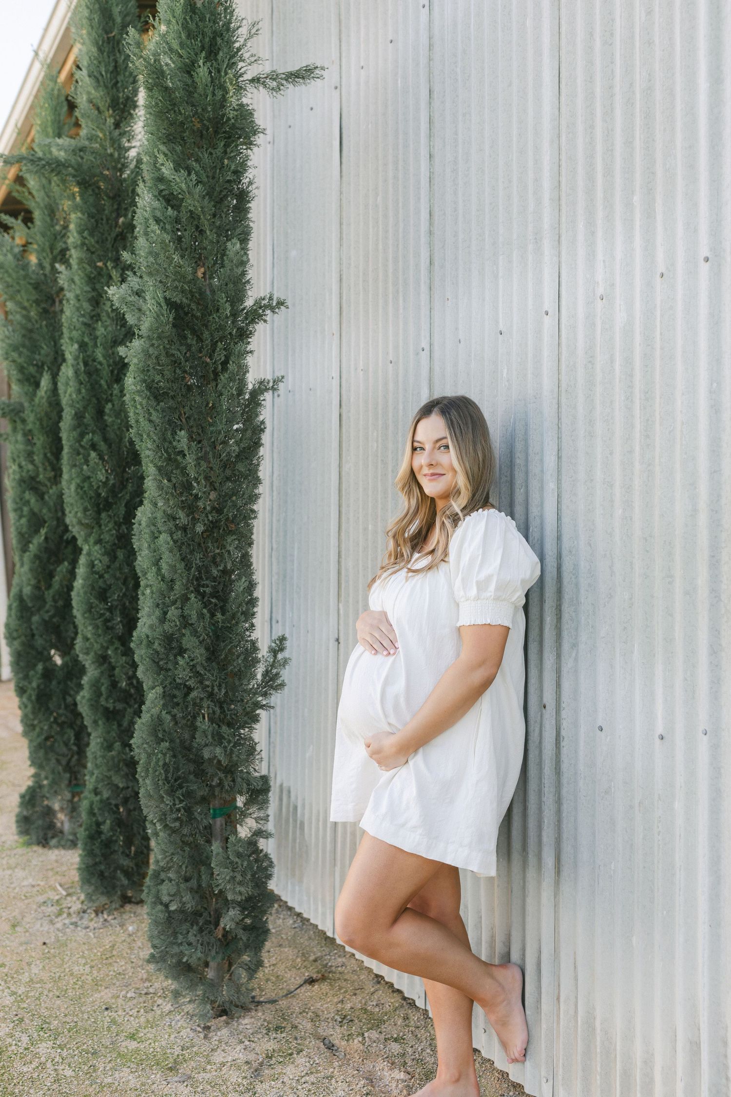 Pregnant woman in white dress leaning against corrugated metal wall beside tall cypress trees.