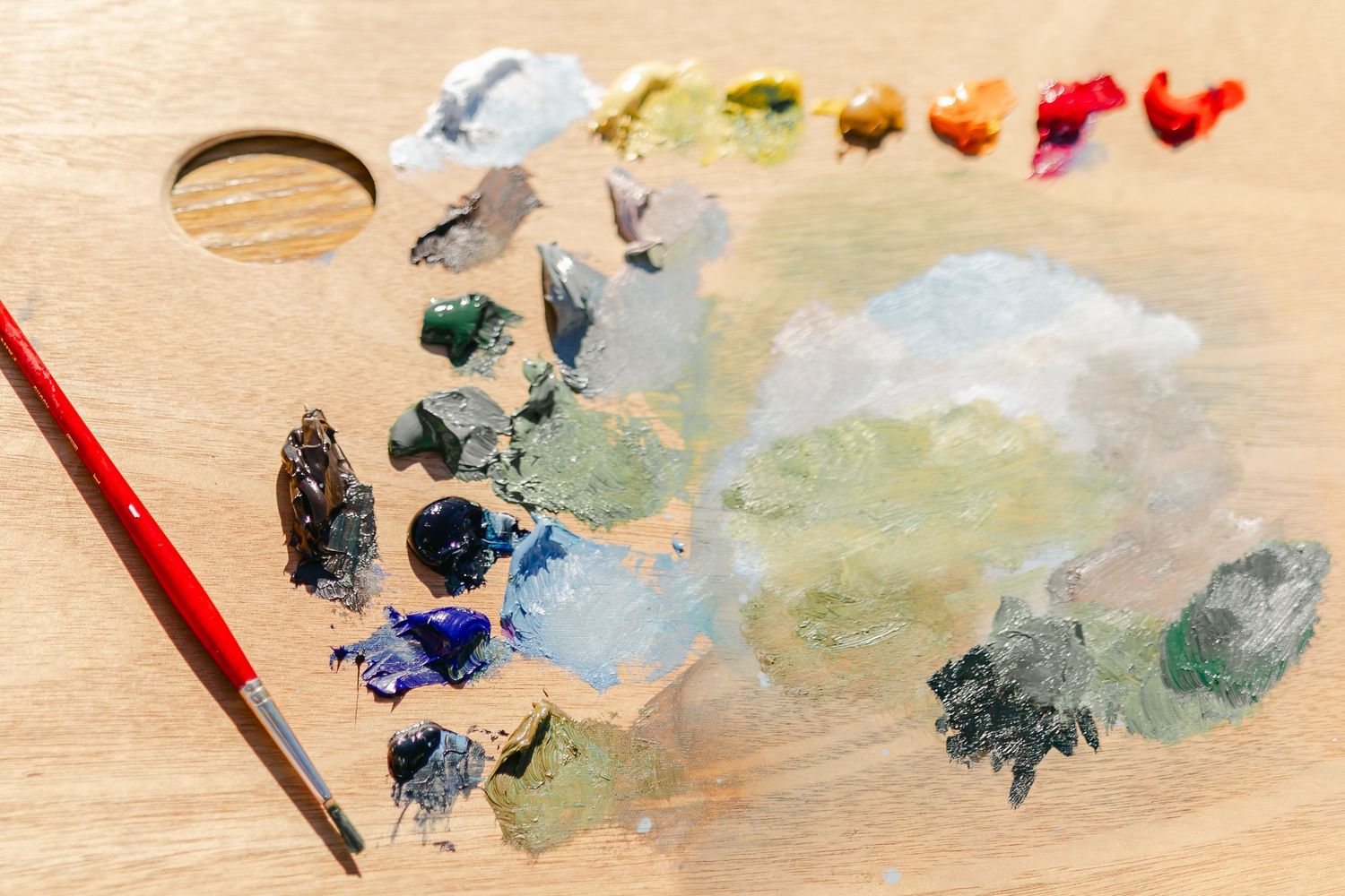 Wooden artist palette with colorful oil paints, mixed pigments, and a red paintbrush for creative painting.