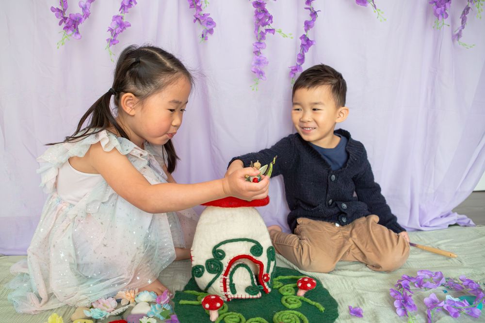 Children work together on a crafting project with decorative purple flowers hanging in the background.