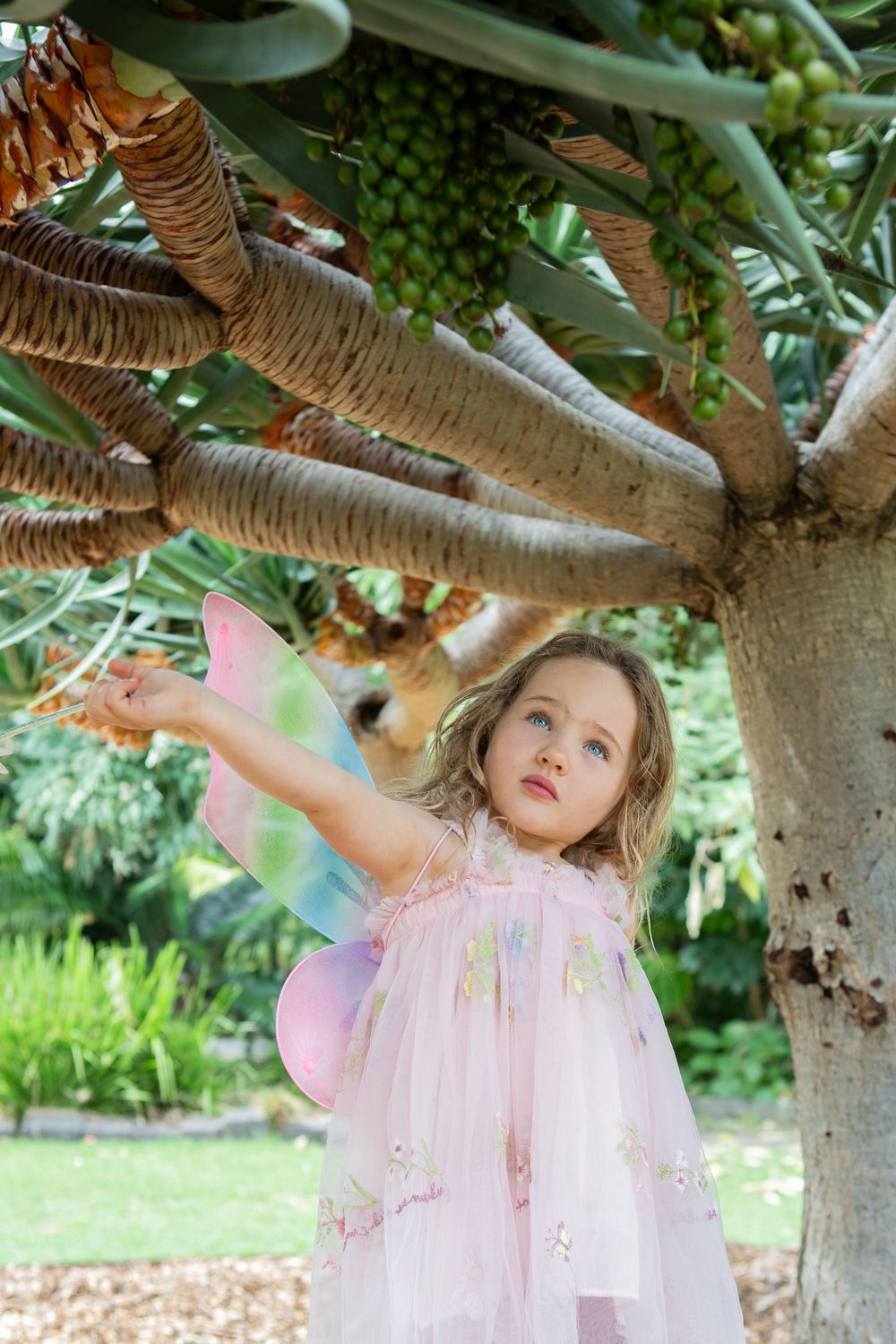 A child in a pink fairy dress with wings plays under a tree branch in a garden setting.