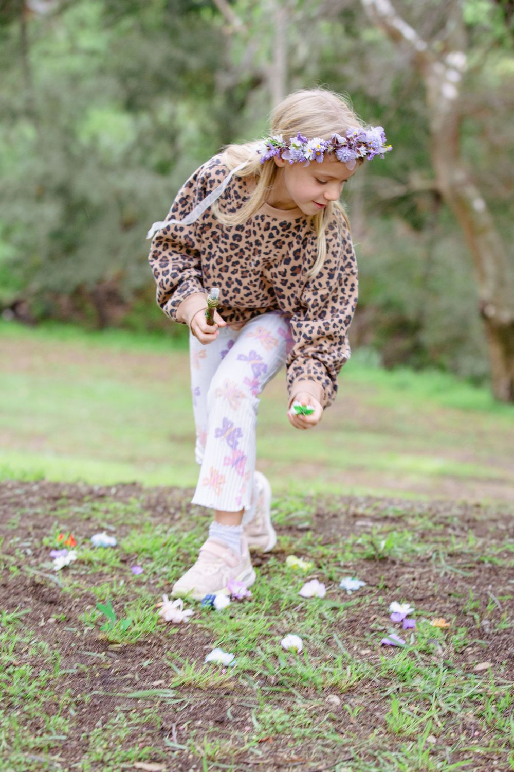 Young girl in leopard print sweater and floral crown picks up Easter eggs scattered on ground in outdoor garden setting.