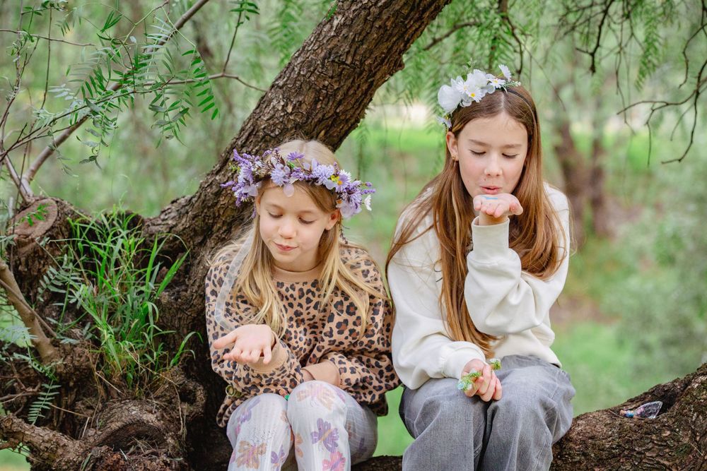 Two girls wearing floral crowns sit on a tree branch, blowing kisses in a lush green outdoor setting.