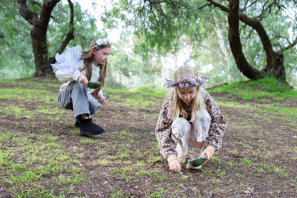 Two girls dressed as fairies crouch in a woodland clearing, wearing wings and floral crowns among mossy trees.