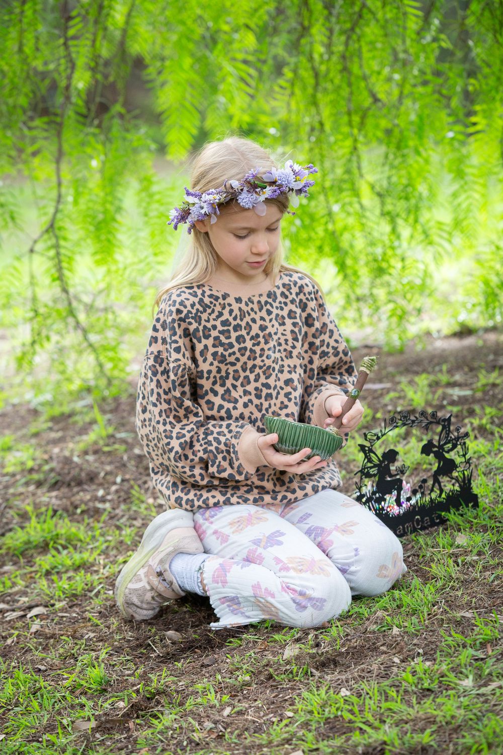 Young girl in leopard print top and floral crown sits cross-legged outdoors, examining small decorative items in a garden.