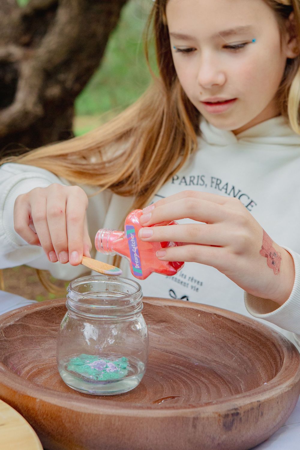 Young girl pouring glitter into a glass jar outdoors, crafting at a wooden table in a garden setting.
