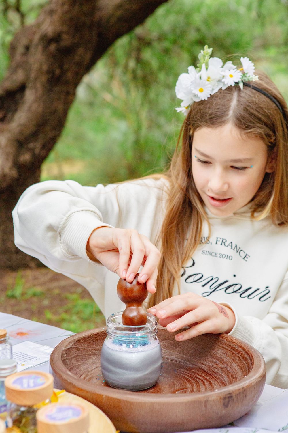 Young girl with flower crown crafting outdoors, pressing clay on pottery wheel at a nature table.