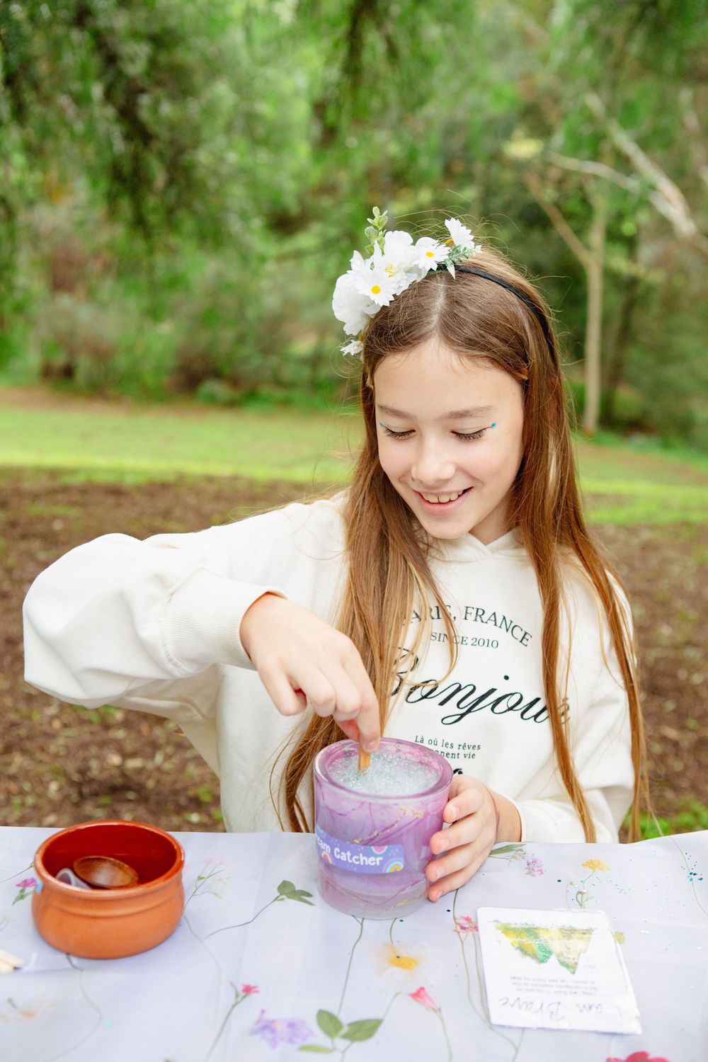 Smiling girl with flower headband stirring a purple mug outdoors at a decorated table.
