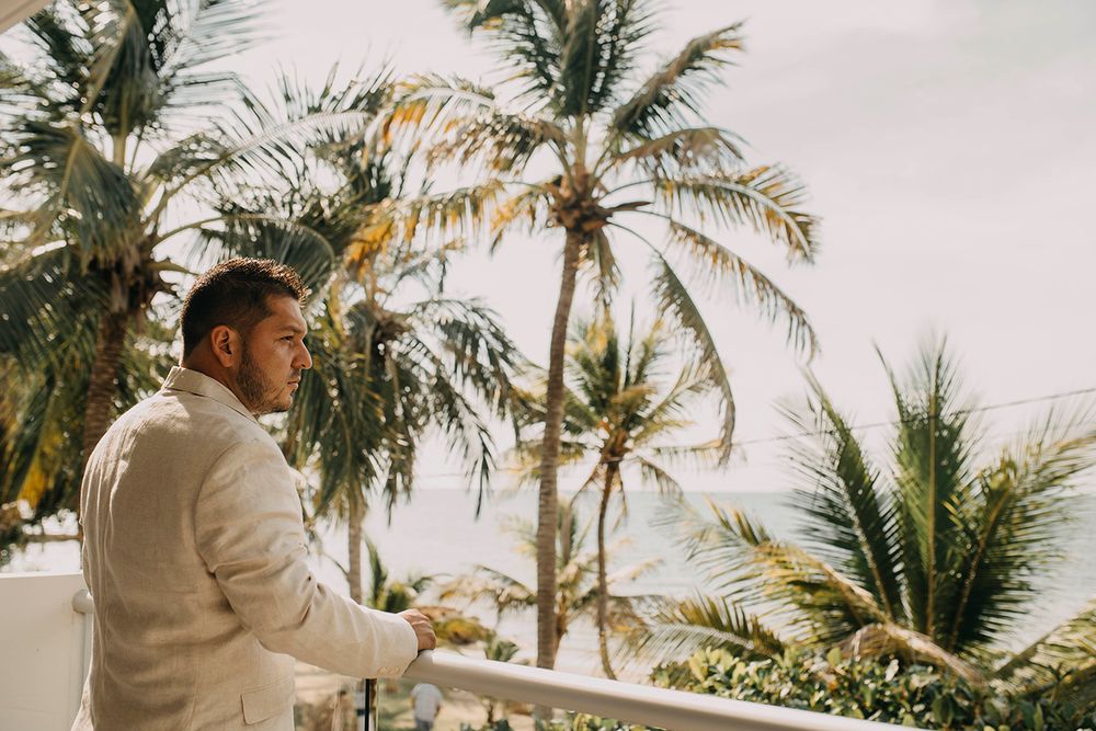 Groom in a white suit standing on a balcony with palm trees and ocean view behind him.