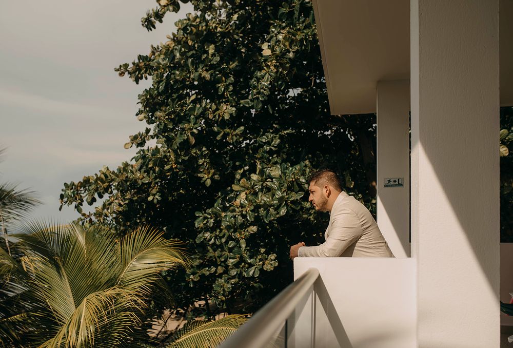 Groom in a beige suit leaning on a white balcony railing surrounded by lush tropical trees.
