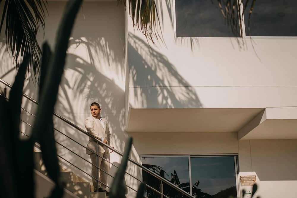 Man standing on a modern balcony surrounded by palm tree shadows on a white building facade.