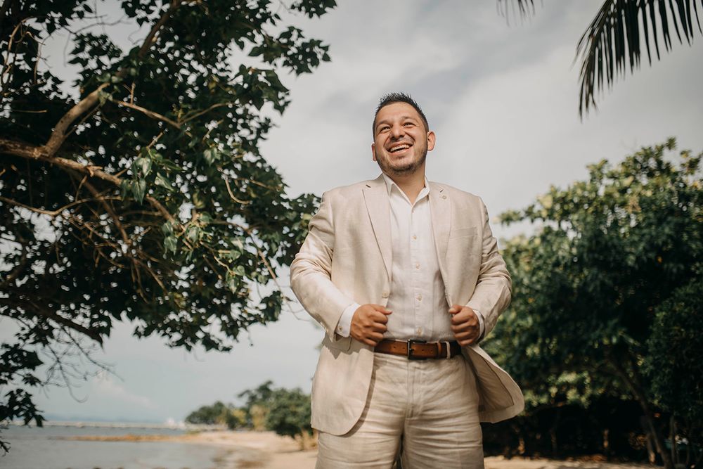 Smiling groom in beige linen suit standing on tropical beach with palm trees and ocean in background.