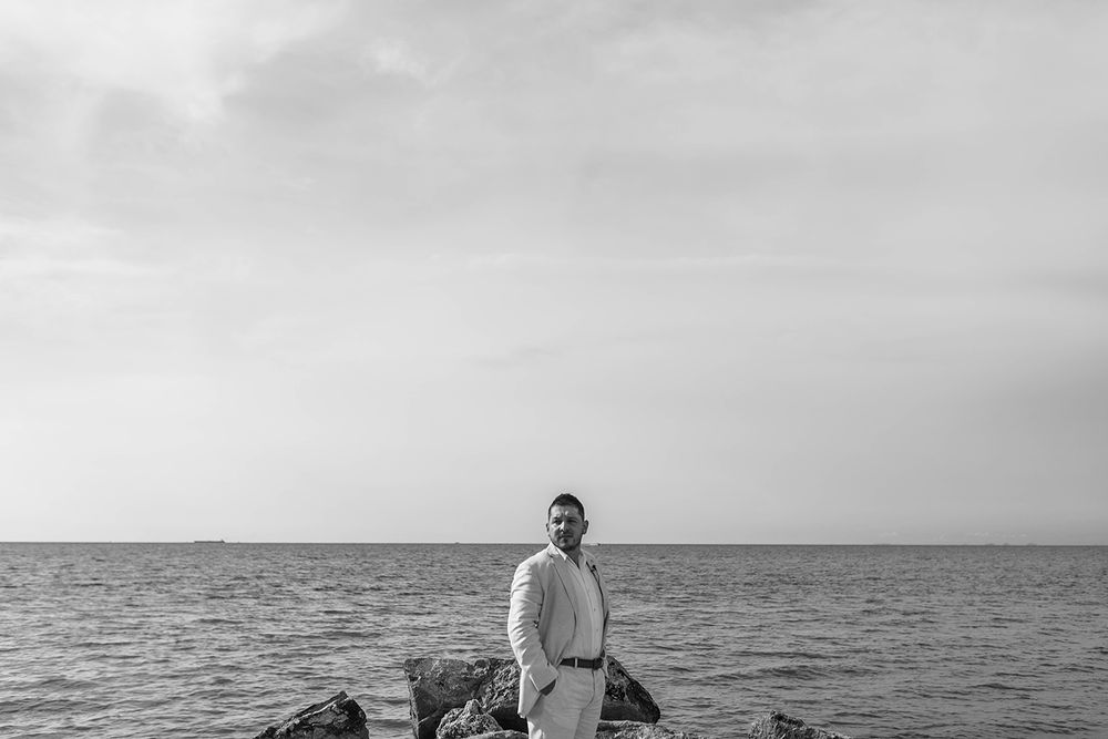 Black and white photo of a man in white outfit standing on rocks by the ocean under a cloudy sky.