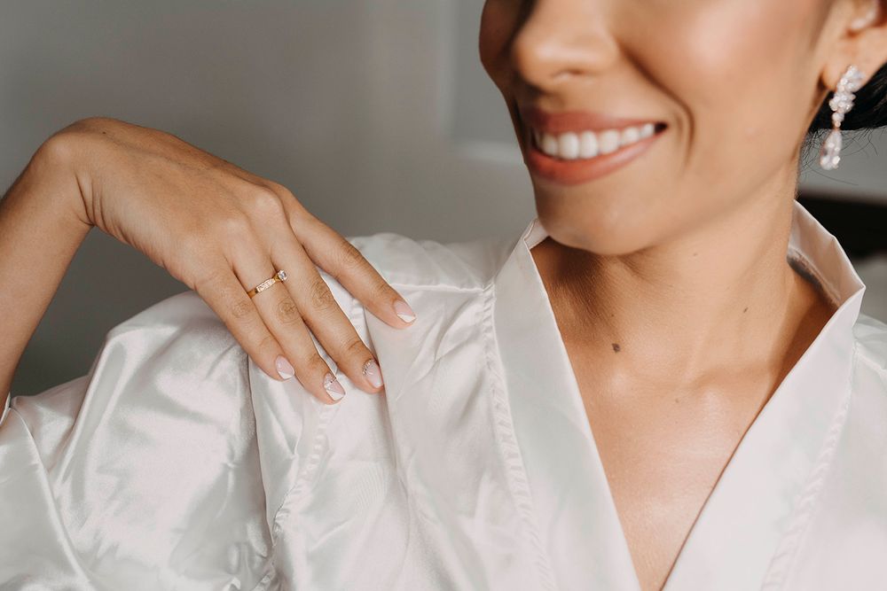 Smiling bride in a white satin robe wearing pearl earrings and a delicate gold engagement ring.