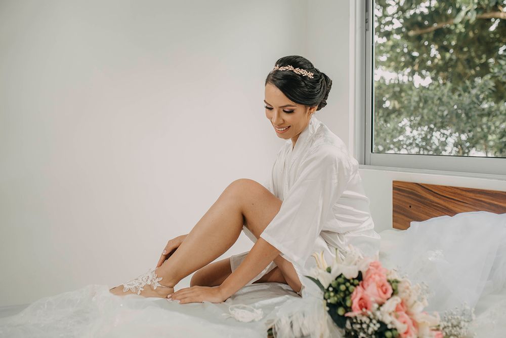 Bride in a white robe sitting on a bed with a floral headpiece and pink bouquet nearby.