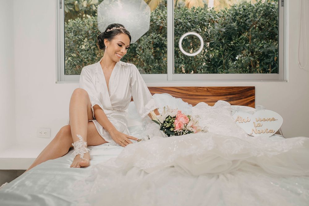 Bride in white satin robe sitting on bed with wedding bouquet and dress, smiling softly in bright room.